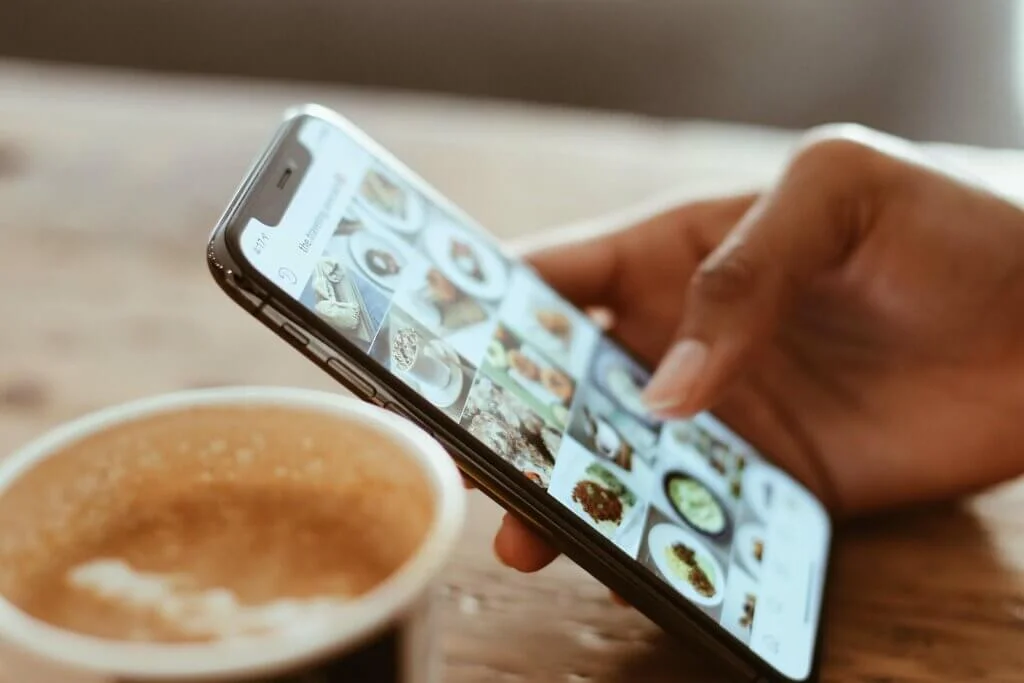 Close-up of a woman's hand holding a phone and scrolling through Instagram. You can see a coffee cup in the background.