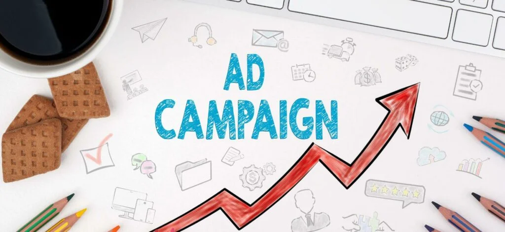 A drawing of a line chart with the word "Ad campaign" in bold, surrounded by small doodles. A keyboard, coffee mug, some biscuits and colored pencils are in the frame.