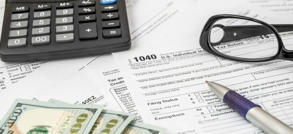 This image features some tax papers sprawled out, a calculator, a pair of glasses, some cash, and a pen.