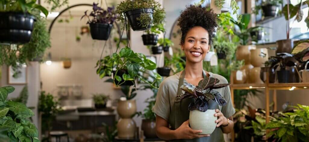 A woman wearing an apron in a small business that sells plants. She is smiling at the camera while holding a potted plant.