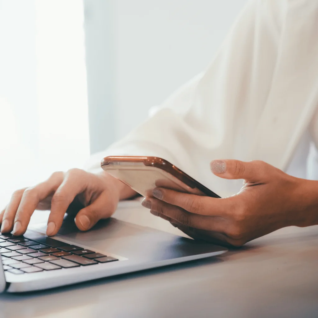 a closeup photo of someone holding their phone with one hand and scrolling through their computer with the other