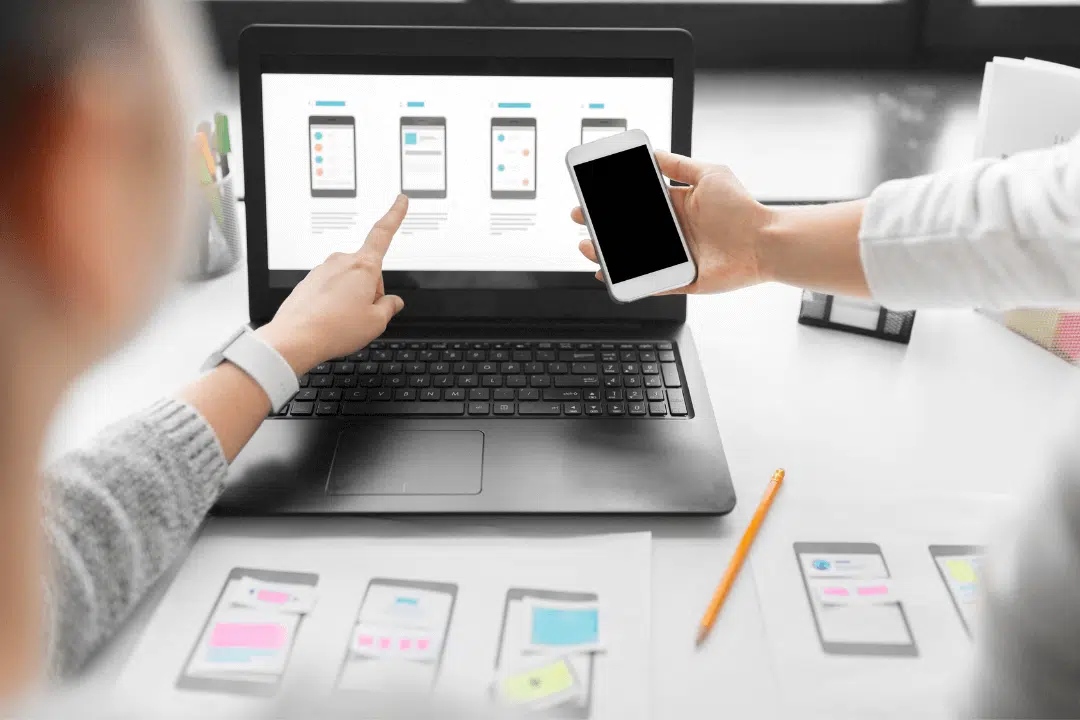 A person pointing at a laptop with web design layouts on it while another is holding a phone in front of the laptop